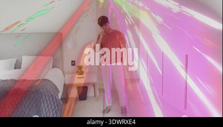 Standing man holding smartphone to ear, glancing down in loft, wearing tee, colored lights over bed Stock Photo