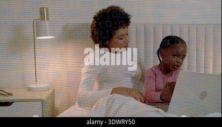 Sitting mother wearing white top and daughter wearing pink top using laptop on bed, with lamp Stock Photo