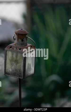 Rusty vintage metal lantern with frosted glass panels on garden stake, set against soft green background with shallow depth of field. Stock Photo