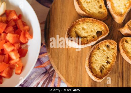 A close-up shot of fresh diced red tomatoes in a white bowl next to toasted artisan bread slices on a wooden board, promoting healthy eating habits an Stock Photo
