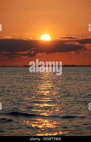 Beautiful golden sunset illuminating the horizon and reflecting on the calm surface of the ocean or sea. The sun, partially hidden behind dark clouds, Stock Photo