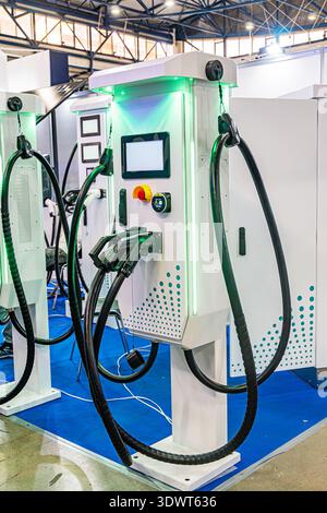 Indoor scene of two electric charging stations, white in color with blue bases and digital interfaces Blue glow emanates from the bases Coiled chargin Stock Photo