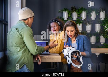 Diverse coworkers testing VR headset and discussing design at counter with laptop, wearing beanie Stock Photo
