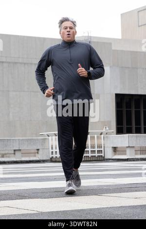 Male middle-aged runner wearing dark clothes and smartwatch running toward camera on striped plaza Stock Photo