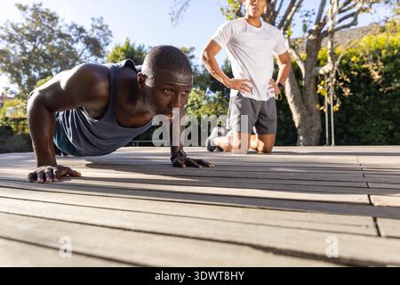 Diverse male training partners doing push-ups on wooden deck wearing fitness tracker, copy space. Outdoor, fitness, motivation, healthy, activewear, training, nature || Model released Stock Photo