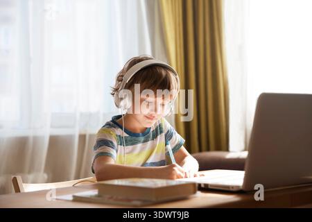child boy in earphones is using a laptop and study online at home. homeschooling, distant learning || Model released Stock Photo
