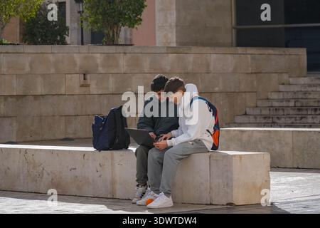 Two male students or friends look at a notebook computer screen while sitting on concrete blocks in a public area in Valencia, Spain. Digital collabor Stock Photo