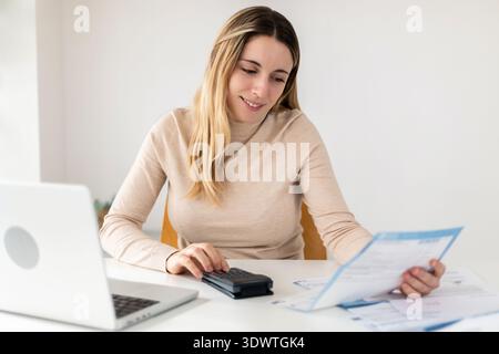 Young adult businesswoman using calculator while checking bills at desk. Stock Photo