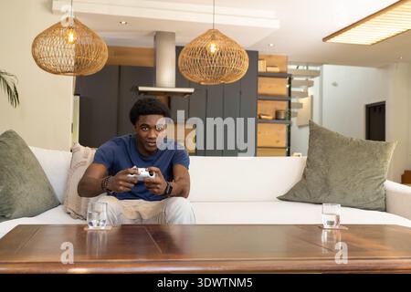 Adult African American male sitting on sofa holding game controller, leaning near table at home. Gamer, livingroom, modern, wooden, warm, cozy, ambien Stock Photo