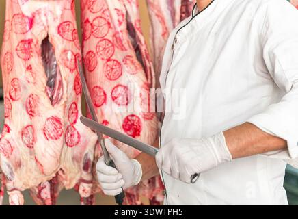 Close up of a professional butcher in uniform and gloves carving fresh raw beef carcass hanging in a slaughterhouse Stock Photo