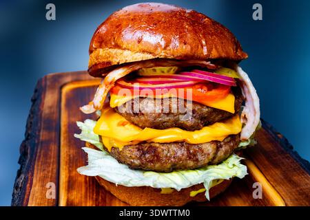 A burger with two beef patties, melted cheese, lettuce, and vegetables sits on a wooden board. It is in a restaurant with blue lighting. Stock Photo