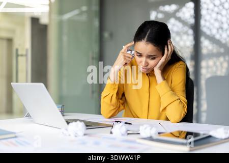Young woman feeling overwhelmed and stressed from work, holding crumpled paper while sitting at her desk with a laptop and documents, experiencing burnout and frustration Stock Photo