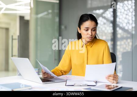Young businesswoman with dark hair in a yellow shirt reviewing financial reports and paperwork at her desk in a modern office, focused and thoughtful on strategy and planning Stock Photo