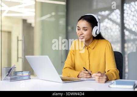Young professional woman wearing a yellow shirt and white headset, smiling and writing notes while working on her laptop in a modern office environment Stock Photo
