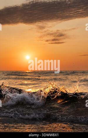 Ocean wave splashing at sunset with golden sunlight reflection on water and dramatic sky background Stock Photo