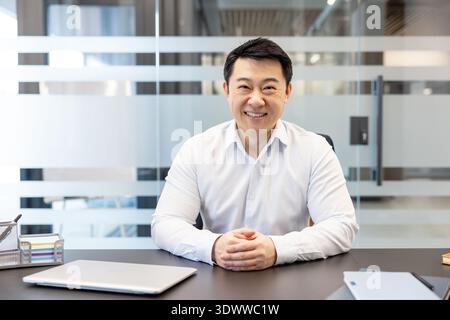 Confident asian businessman smiling at the camera, sitting at his office desk in a professional modern co-working space, representing success, leadership, and a positive attitude Stock Photo