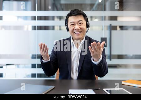 Asian businessman, smiling and making hand gestures, wearing a headset, participating in an online video conference in a modern office, representing customer support and remote work Stock Photo