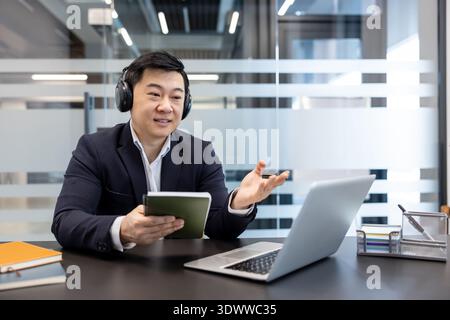 Asian businessman wearing headphones participating in an online video call meeting, talking and gesturing while holding a notebook, working remotely from a modern office Stock Photo