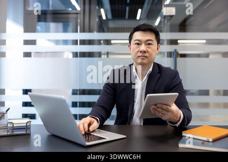 Young asian businessman in suit multitasking at office desk, looking at camera while using laptop and holding a tablet, focused on digital workflow and productivity Stock Photo