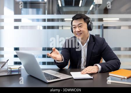 Asian businessman in headphones working at laptop having online video call, communicating and gesturing in modern office while involved in remote meeting or webinar Stock Photo