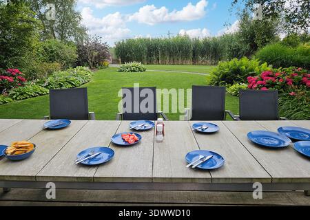 Wooden outdoor dining table set with blue plates and cutlery on terrace overlooking green landscaped garden lawn, Outdoor dining, Summer gathering Stock Photo