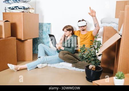 Couple sitting among cardboard boxes while using VR headset and tablet for visualization their future interior and home, Interior planning, renovation Stock Photo