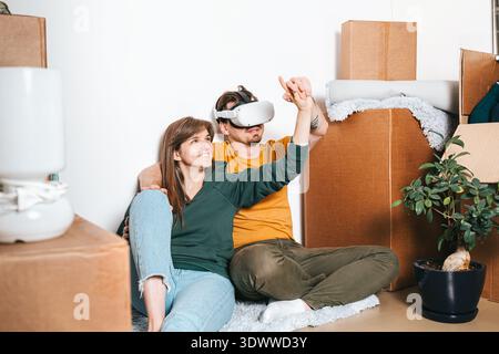 Man wearing virtual reality headset and woman pointing while sitting in room with packed cardboard boxes, renovation, interior planning and visualizat Stock Photo