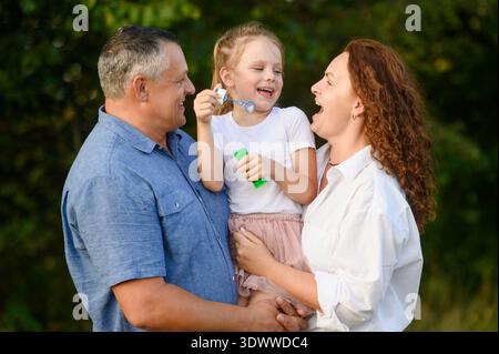 Mother and father hold baby girl, laughing happily while blowing bubbles on summer day, close-up. Family vacation. Family fun concept. International Day of Families Stock Photo