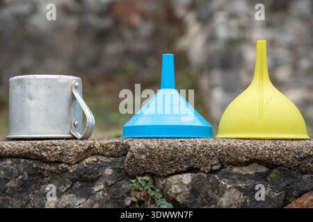 Metal mug, plastic funnels in blue, yellow standing on stone surface outdoors, Household utensils, simple objects suitable for product presentation Stock Photo