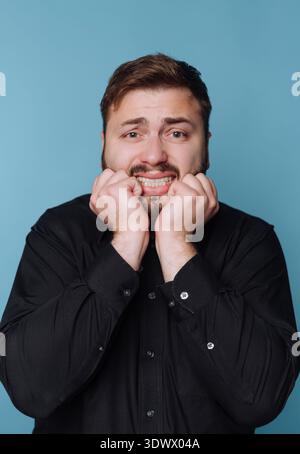 A young man with short hair shows a nervous expression, biting his lip and clasping his hands together, surrounded by a calming blue backdrop. His eye Stock Photo