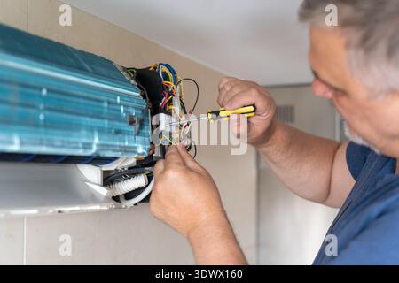 A technician is focused on repairing an air conditioning unit by adjusting wires and screws. The work is taking place indoors, highlighting attention Stock Photo