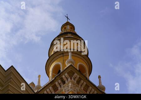 Low-angle view of yellow Orthodox church tower with ornate dome and cross against blue sky.Menton,France Stock Photo