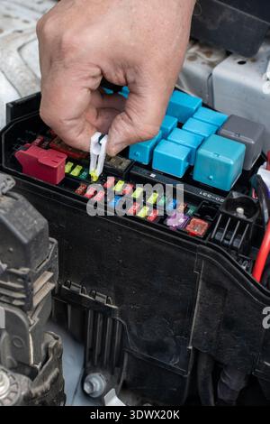 Various colored relays and fuses are arranged in the fuse box, indicating a maintenance task underway. A person is focused on replacing fuses under th Stock Photo