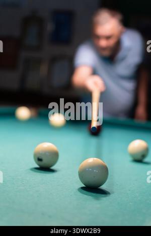 Concentrated mature man playing billiards, carefully aiming his cue stick to hit the white ball on a green billiard table in a dimly lit game room, sh Stock Photo