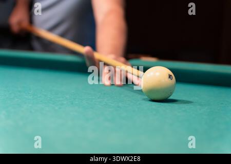 Concentrated mature man playing billiards, carefully aiming his cue stick to hit the white ball on a green billiard table in a dimly lit game room, sh Stock Photo