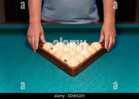 Billiard player arranging numbered white balls with a wooden triangle rack on a vibrant green pool table, focusing intently on setting up for an excit Stock Photo
