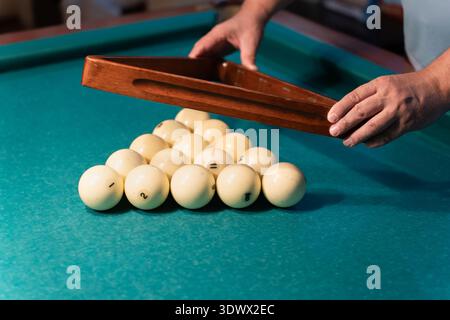 Billiard player arranging numbered white balls with a wooden triangle rack on a vibrant green pool table, focusing intently on setting up for an excit Stock Photo