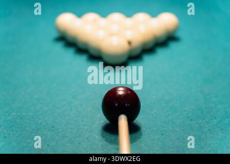 Concentrated man playing billiards, carefully aiming his cue stick to hit the white ball on a green billiard table in a dimly lit game room, showcasin Stock Photo