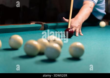 Concentrated mature man playing billiards, carefully aiming his cue stick to hit the white ball on a green billiard table in a dimly lit game room, sh Stock Photo