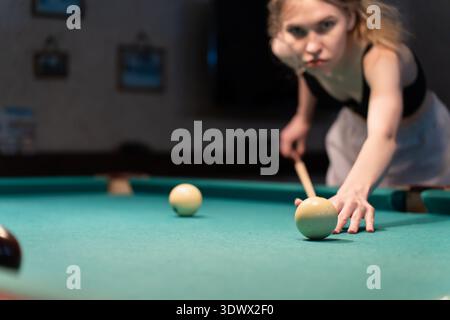 Young caucasian woman playing snooker or billiards aiming the white ball on a green pool table with a wooden cue stick, in a blurred game room Stock Photo