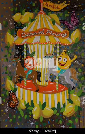 Whimsical carousel painting with smiling fruit characters and butterflies on striped platform.Menton,France Stock Photo