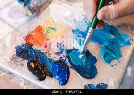A hand holds a brush, mixing vibrant blue paint among a palette of colors in a cozy artist's studio. The table features splashes of orange, black, and Stock Photo