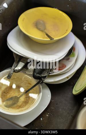 Dishes are piled high in a kitchen sink, featuring bowls, plates, and utensils covered in food residue. The setting reveals a busy kitchen atmosphere, Stock Photo