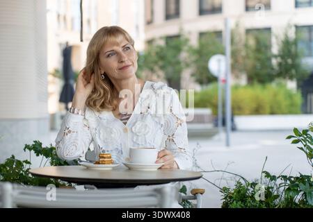 Portrait of a mature woman enjoying a personal moment at a cafe table, framed by soft light and city calm. Stock Photo