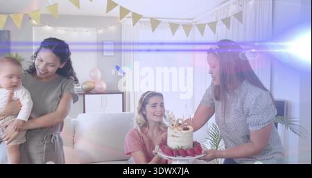 Presenting woman bringing cake with candles and strawberries to woman in pink top on sofa, balloons Stock Photo