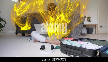 Sitting male typing on laptop on floor in bedroom, yellow particles rising, open suitcase Stock Photo