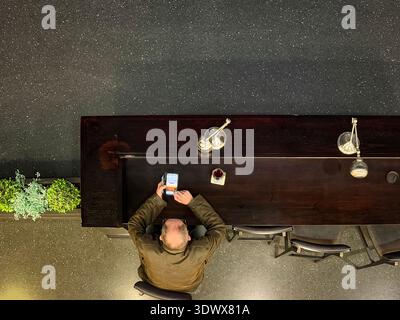 Overhead view of a man seated at a dark wooden bar or counter looking at his smartphone glasshouse concert venue uk Stock Photo