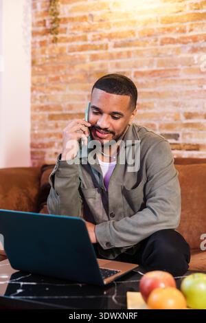 Young man working from home talking on phone and using laptop Stock Photo