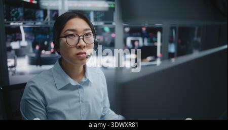 Young Asian Specialist in Crisp Blue Shirt, Focused on Real Time Analytical Dashboards. Woman Wearing Professional Headset For Instant Communication. New Generation Experts in Investment Environment. Stock Photo