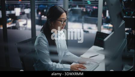 Asian Female Financial Specialist Sits at Innovative Workspace, Eyes Scanning Multiple Displays Filled With Trading Indices. Young Lady Actively Uses Keyboard to Input Data and Execute Market Orders. Stock Photo